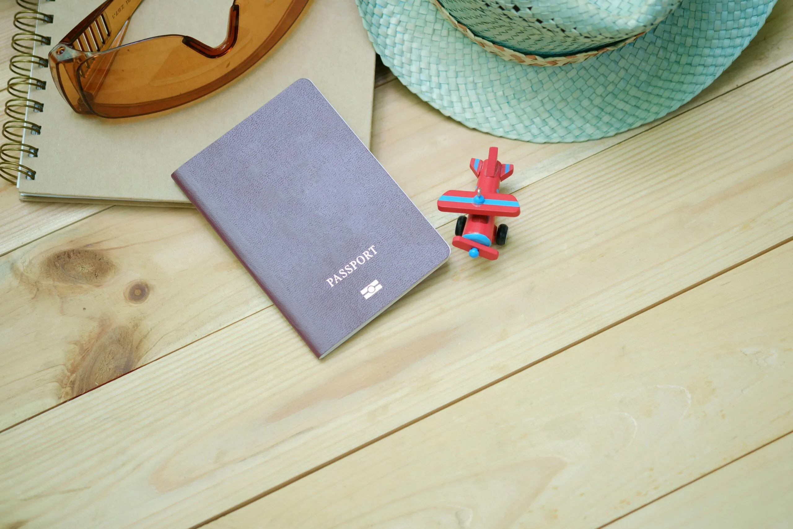 Flat lay of travel essentials including passport, notebook, toy airplane, sunglasses, and straw hat.