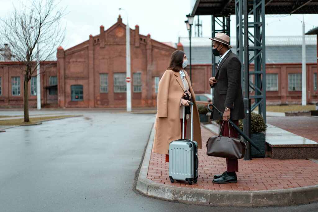 A fashionable couple in masks stands with luggage outside a historic building.